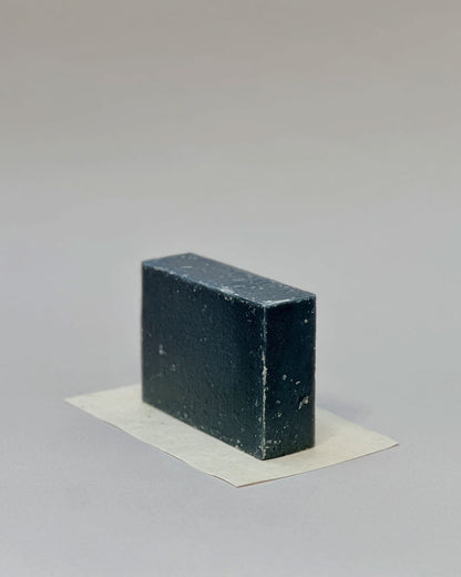 A rectangular bar of charcoal soap sitting on its side on a rectangular piece of white paper against a seamless, light grey background. 
