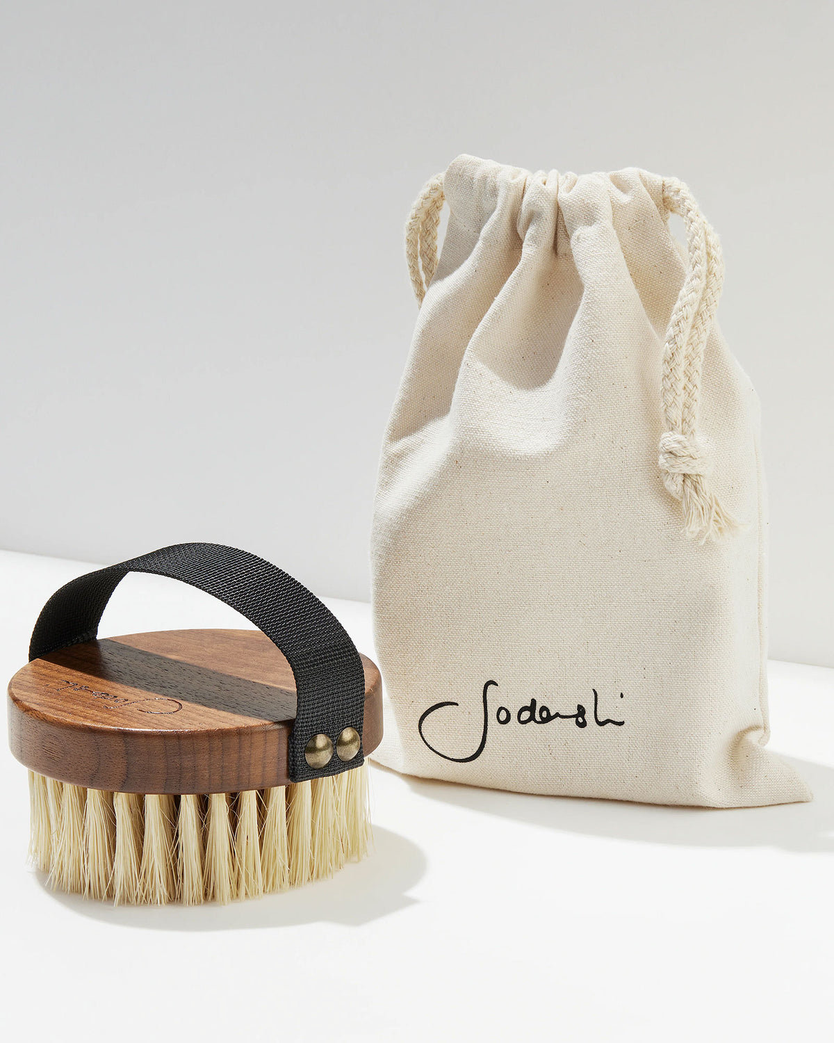 A Sodashi Dry Body Brush and a Sodashi branded, cream-coloured drawstring pouch sit on a white surface with white background. 