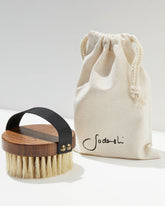 A Sodashi Dry Body Brush and a Sodashi branded, cream-coloured drawstring pouch sit on a white surface with white background. 