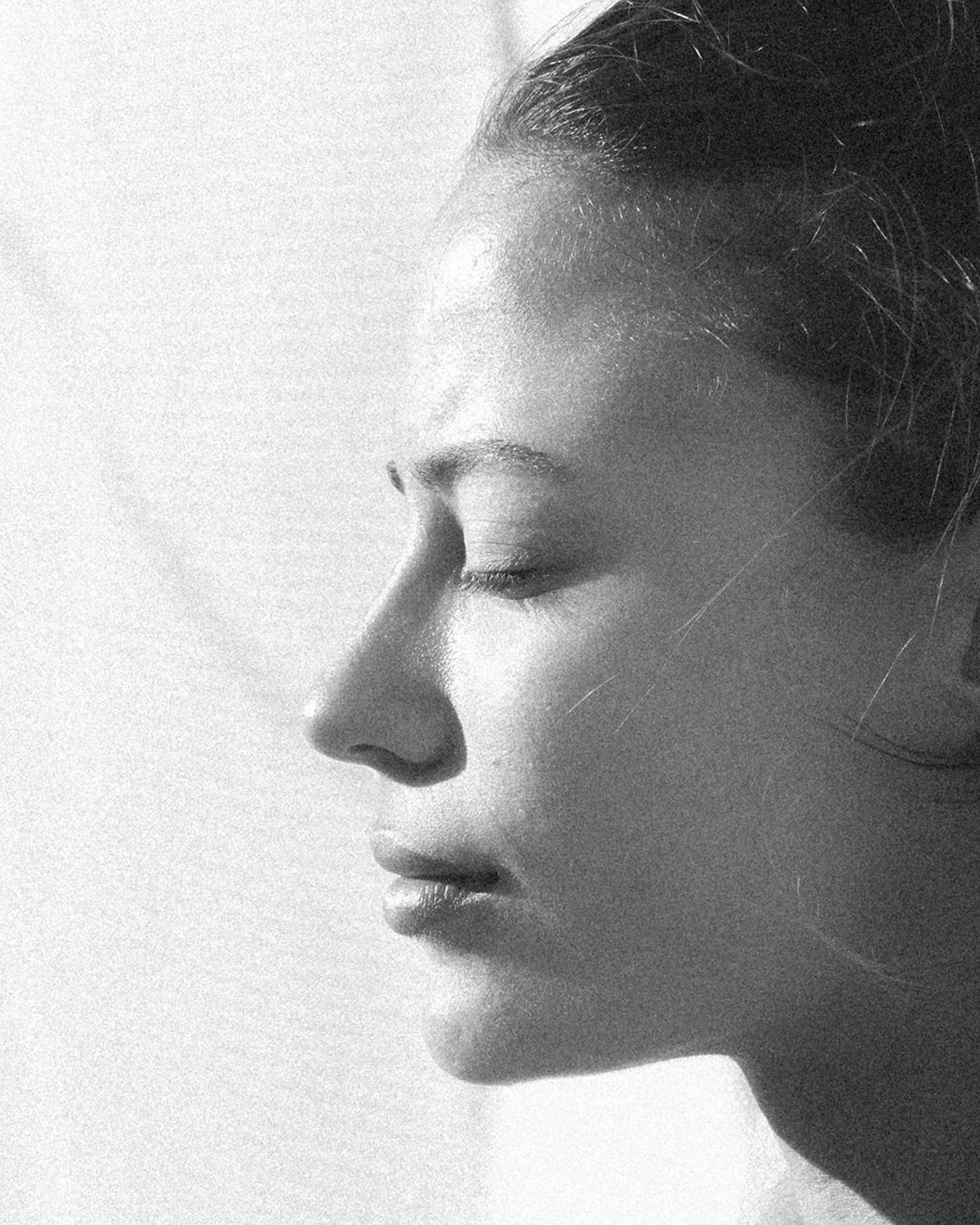Black and white close-up portrait of a woman in profile with her eyes closed, shown against a bright light background.