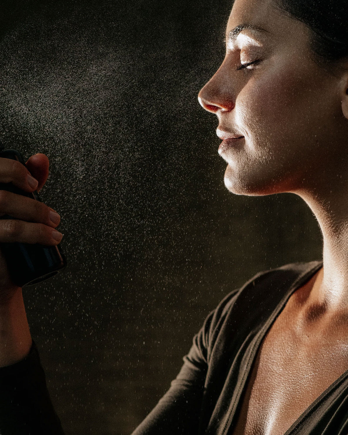 A close-up side profile of a woman holding and spraying a dark mist bottle, with fine droplets visible against a dark brown background.