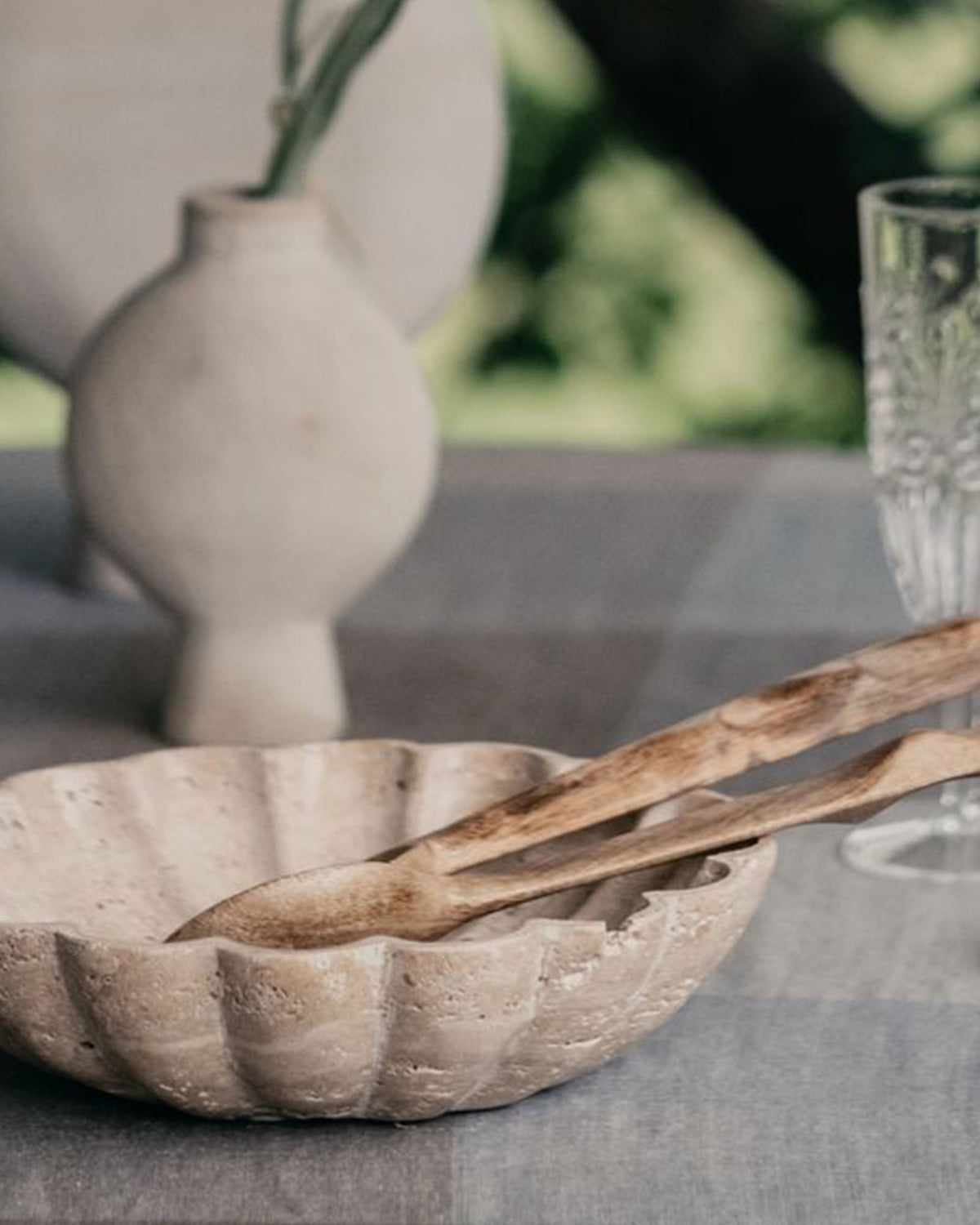 A scalloped travertine serving bowl with wooden serving spoons on a light grey linen tablecloth next to a white vase and drinking glass.