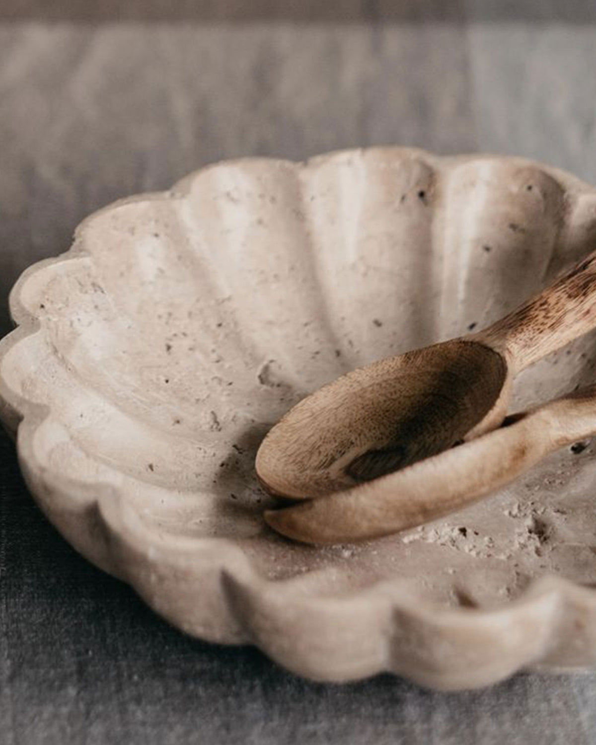 A scalloped travertine serving bowl with wooden serving spoons on a light grey linen tablecloth.