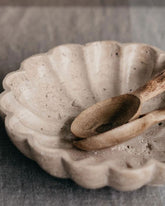 A scalloped travertine serving bowl with wooden serving spoons on a light grey linen tablecloth.