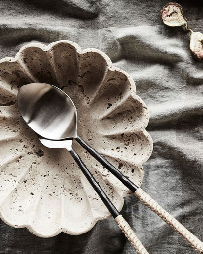 A scalloped travertine serving bowl with woven handled metal serving utensils on a light grey linen tablecloth with a cut fig.