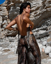A woman stands outdoors against a rocky desert cliff with her back facing, she is tying two ends of a top together behind her back, she is wearing a matching set of a mottled purple wrap top and pants. 