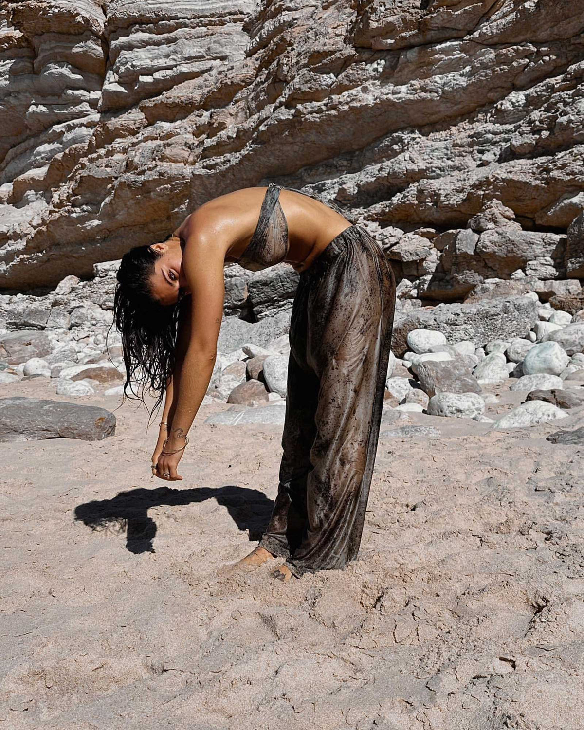 A woman in a sheer, matching set of a mottled purple silk wrap top and silk pants bends forward on a sandy beach with a rocky cliff in the background.