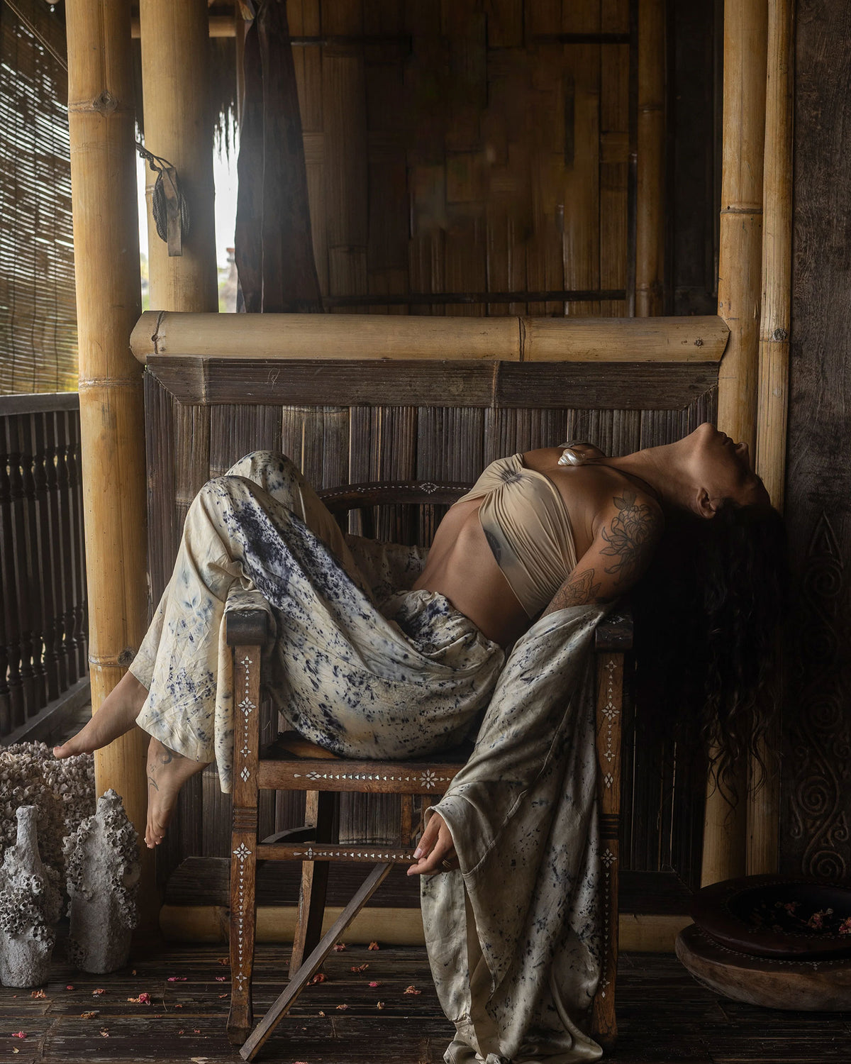 A woman reclines on a wooden chair in a rustic bamboo interior, wearing a beige bandeau-style top and loose patterned silk pants with a draped fabric over her body.
