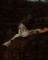 A woman reclines on dark rocks outdoors, wearing a matching silk top and pants in a brown and cream abstract print. The scene is dim and moody, with the background consisting of dark natural rocks and foliage.