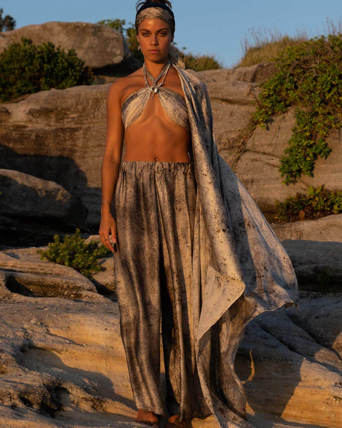 A model stands on rocky outdoor terrain wearing a matching three-piece silk outfit with a halter-style top, loose draped pants, and a flowing head wrap. The background features natural rock formations and sparse greenery under warm sunlight.