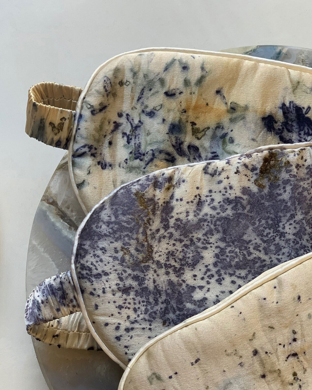 Three padded eye masks with elastic straps, featuring abstract tie-dye and speckled patterns in cream, navy, and brown, arranged on a neutral surface.