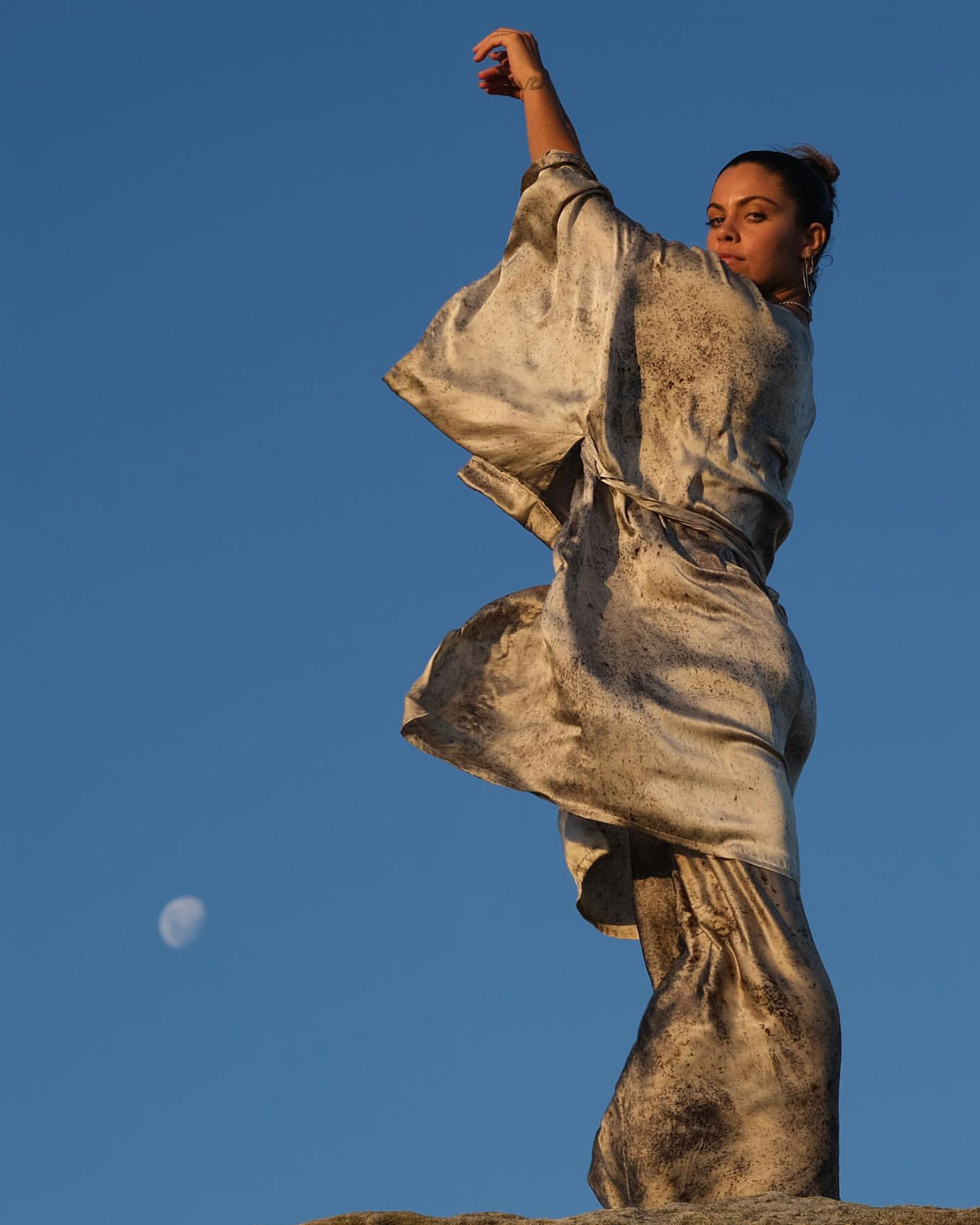 Woman dressed in purple and white coloured silk kimono robe and matching pants stands in wind with arms raised near a semi-full moon and bright blue sky.