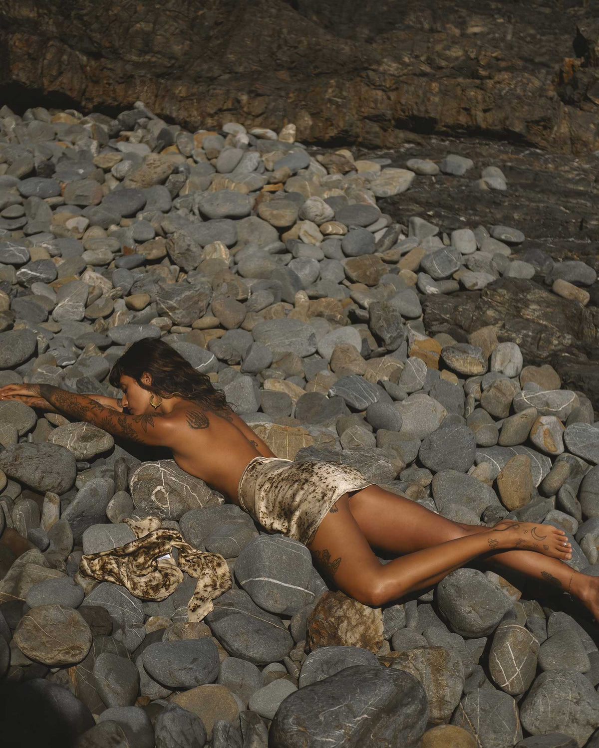 Tattooed woman laying topless on grey river rocks, she has a brown-mottled silk cloth over her buttocks and a silk scarf on the rocks next to her.