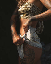 A side profile of a tattooed woman tying a purple and cream mottled silk sarong around her waist, she is wearing a brown-mottled silk bandeau top.