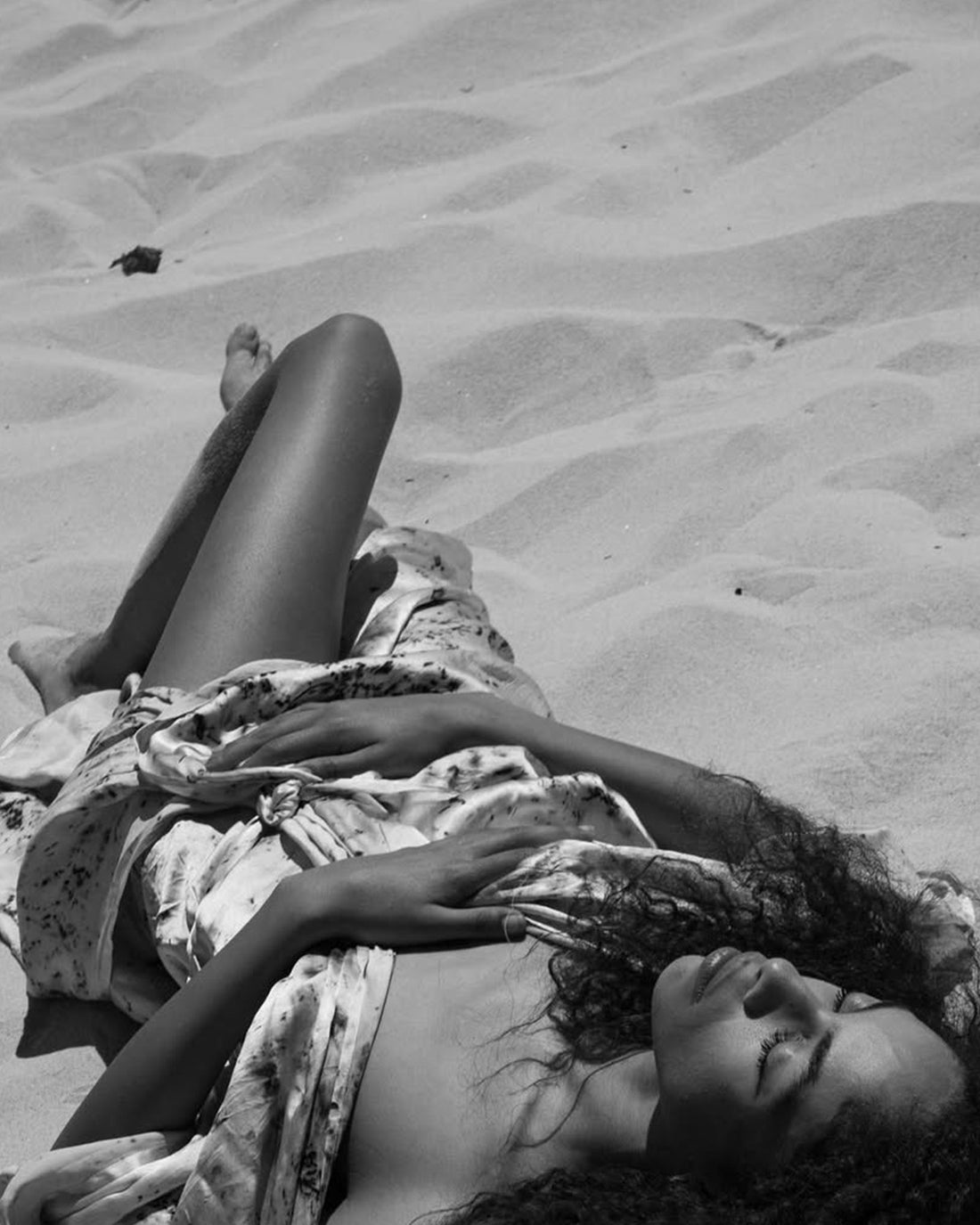 A black and white photograph of a woman lying on sand, wrapped in a silk robe.