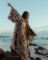 A woman standing amongst rocks along a shoreline, she wears a matching silk robe, top and trouser set. Her arms are stretched and open showing the kimono style sleeves of the robe. 