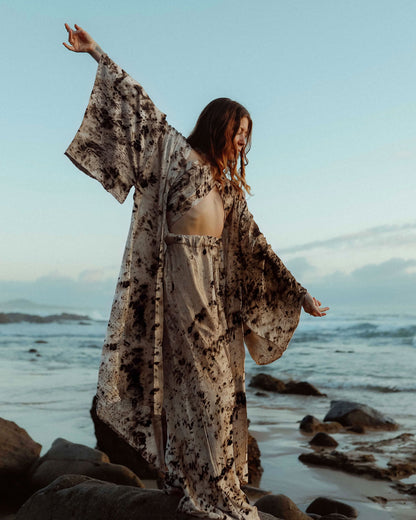 A woman standing amongst rocks along a shoreline, she wears a matching silk robe, top and trouser set. Her arms are stretched and open showing the kimono style sleeves of the robe. 
