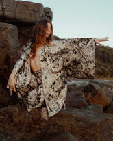 A woman crouching on a brown rock by the ocean wearing a matching silk robe and pants.