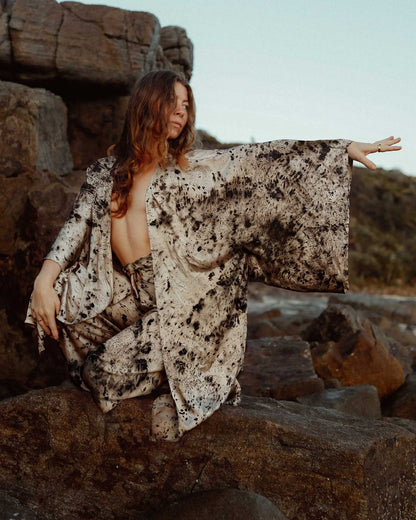 A woman crouching on a brown rock by the ocean wearing a matching silk robe and pants.