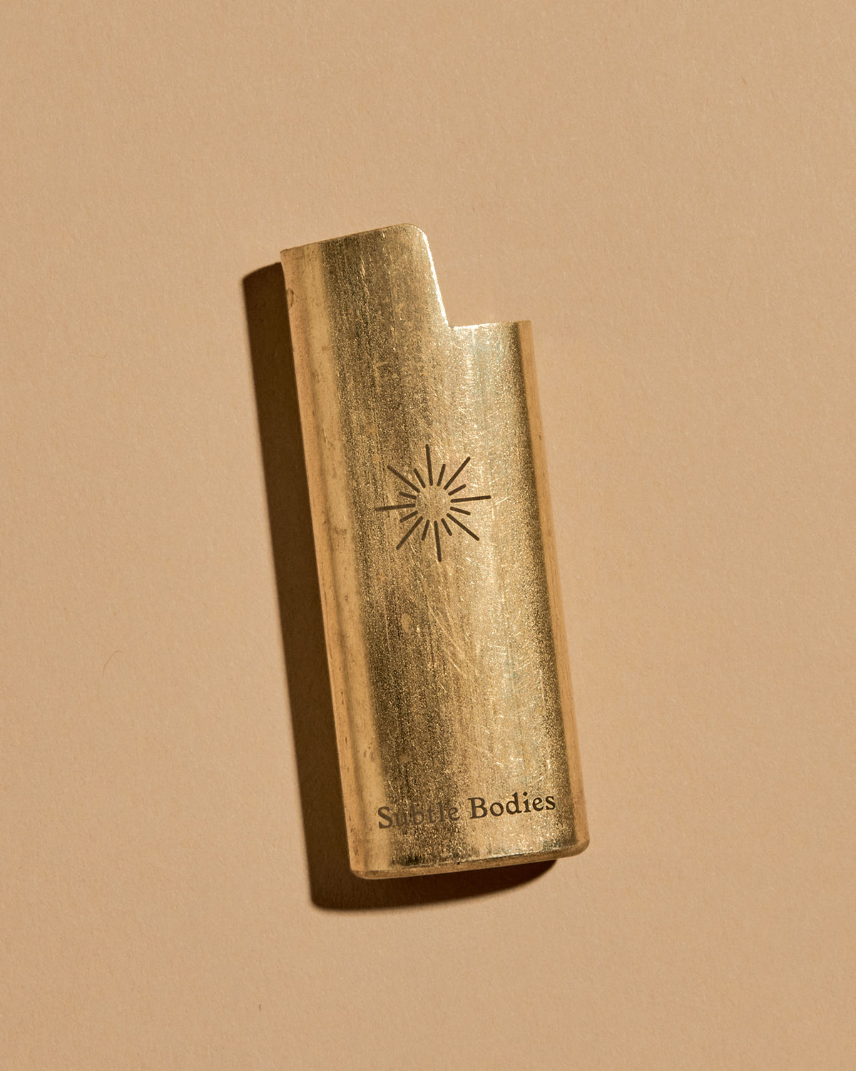 A mini BIC lighter in a brass lighter case with a matte finish. A geometric sun and the Subtle Bodies logo is etched on to the front of the lighter case. Set on a seamless, light orange background. 