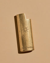 A mini BIC lighter in a brass lighter case with a matte finish. A geometric sun and the Subtle Bodies logo is etched on to the front of the lighter case. Set on a seamless, light orange background. 