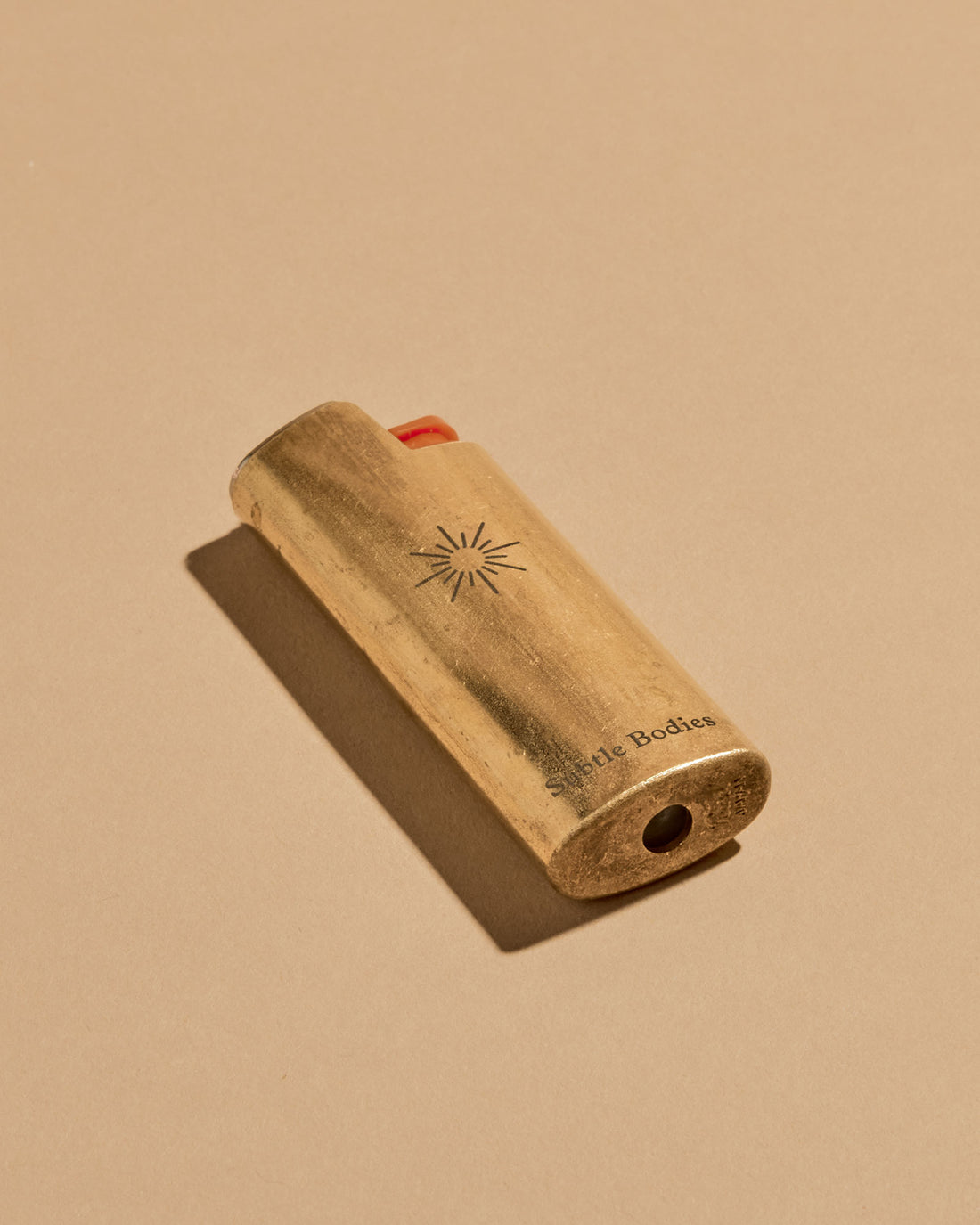 A mini BIC lighter in a brass lighter case with a matte finish. A geometric sun and the Subtle Bodies logo is etched on to the front of the lighter case. Set on a seamless, light orange background. 