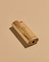 A mini BIC lighter in a brass lighter case with a matte finish. A geometric sun and the Subtle Bodies logo is etched on to the front of the lighter case. Set on a seamless, light orange background. 