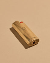 A mini BIC lighter in a brass lighter case with a matte finish. A geometric sun and the Subtle Bodies logo is etched on to the front of the lighter case. Set on a seamless, light orange background. 