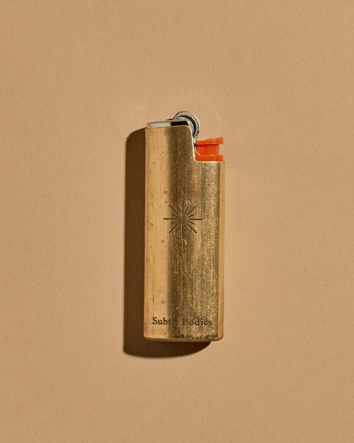 A mini BIC lighter in a brass lighter case with a matte finish. A geometric sun and the Subtle Bodies logo is etched on to the front of the lighter case. Set on a seamless, light orange background. 