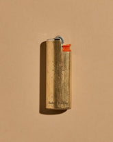 A mini BIC lighter in a brass lighter case with a matte finish. A geometric sun and the Subtle Bodies logo is etched on to the front of the lighter case. Set on a seamless, light orange background. 