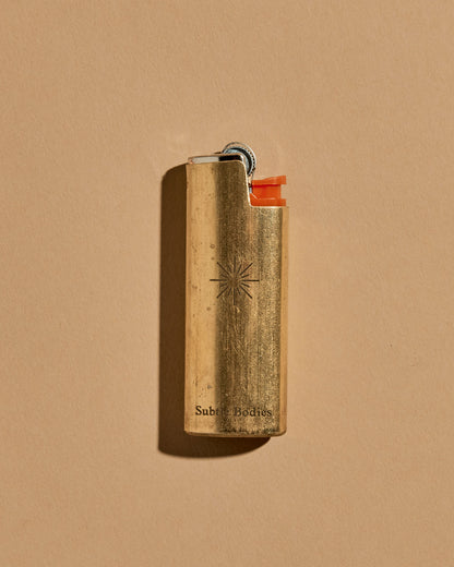 A mini BIC lighter in a brass lighter case with a matte finish. A geometric sun and the Subtle Bodies logo is etched on to the front of the lighter case. Set on a seamless, light orange background. 