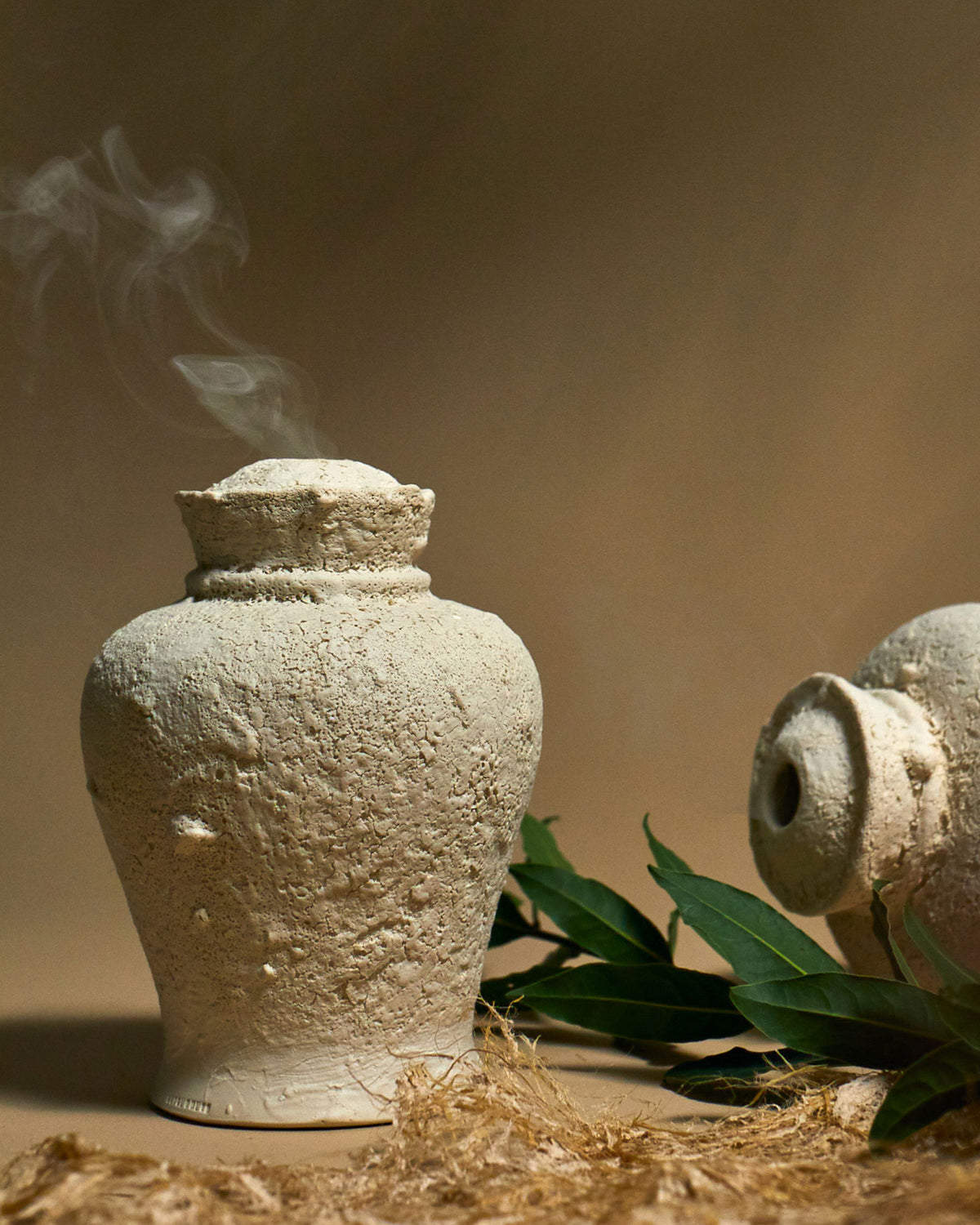 An off-white sculptured ceramic urn with a heavily textured surface sits on a light orange surface that bleeds into the background. Incense smoke smoulders from the top of the urn. A second urn sits to the right on its side with a branch of green leaves.