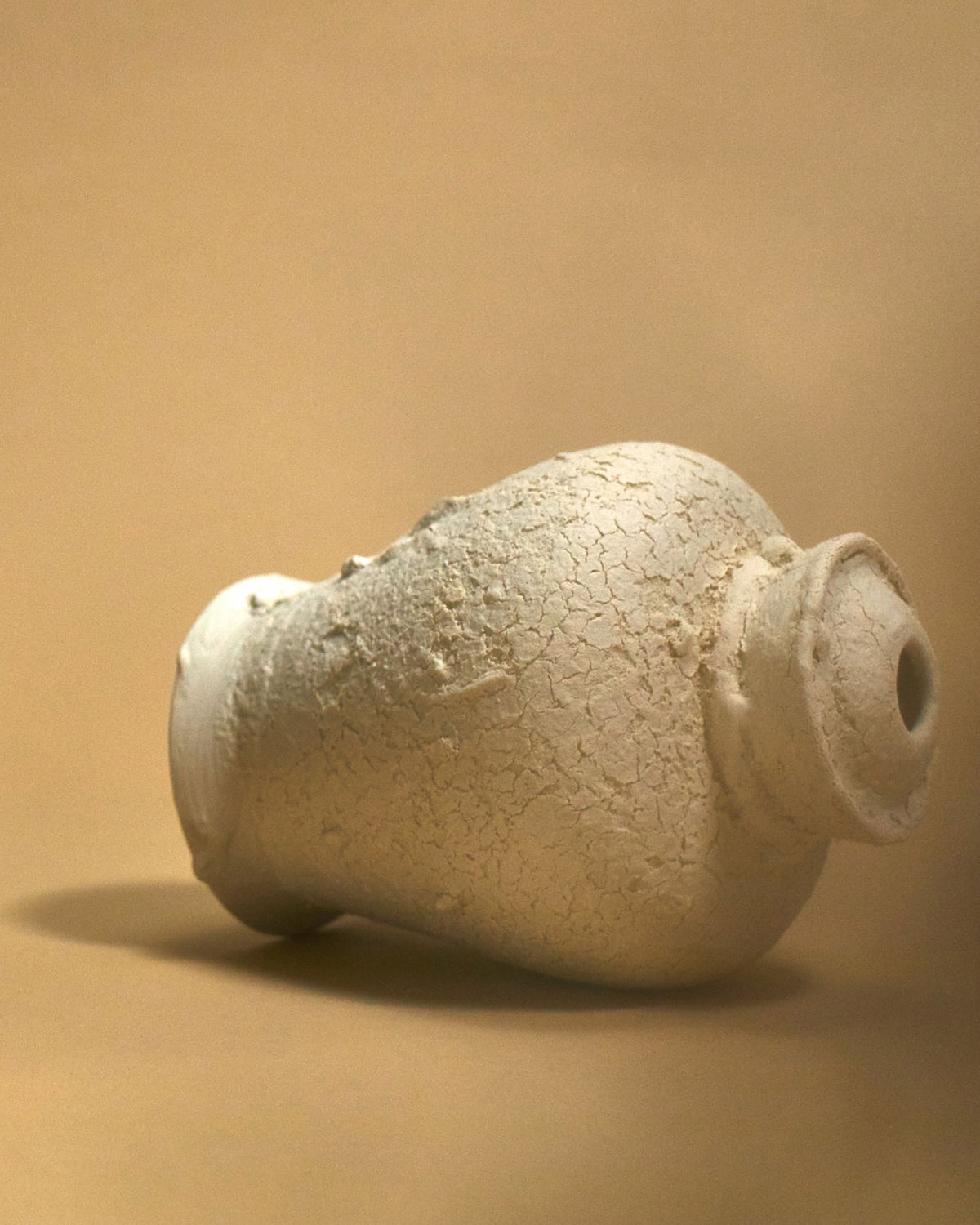 An off-white sculptured ceramic urn with a heavily textured surface lays on its side against a light orange surface and background.