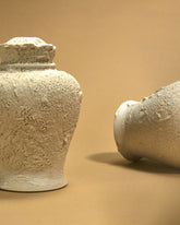An off-white sculptured ceramic urn with a heavily textured surface sits on a light orange surface that bleeds into the background. A second urn sits to the right on its side.