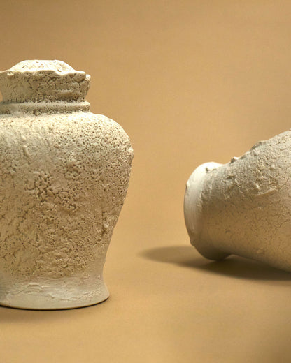 An off-white sculptured ceramic urn with a heavily textured surface sits on a light orange surface that bleeds into the background. A second urn sits to the right on its side.