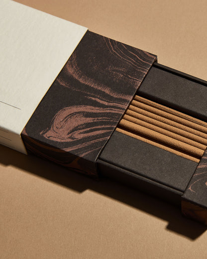 An opened red marbled box of Subtle Bodies Australian Sandalwood Incense showing the contents of light brown incense sticks. Set on a light orange background.