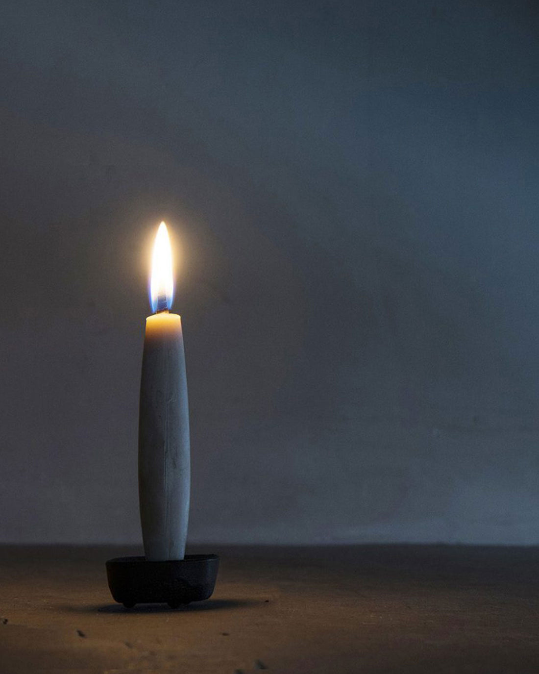 A single lit taper candle upon a black iron holder, standing on a surface against a dim, cool-toned background.