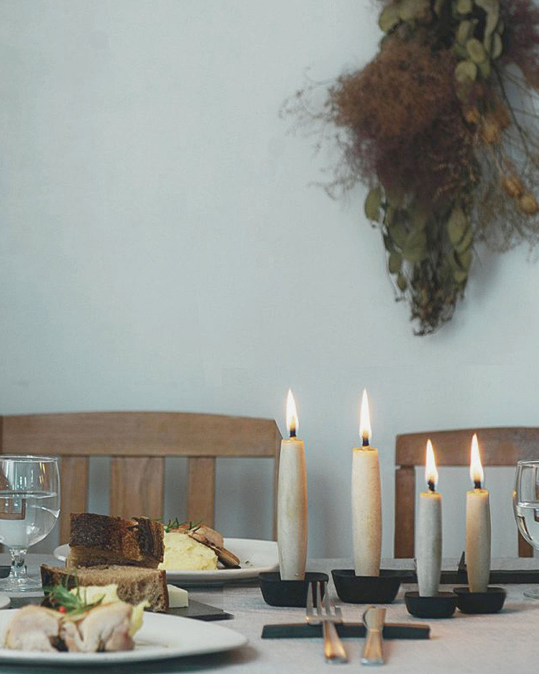 A set dinner table with dinner plates containing bread, vegetables and meat. Silver cutlery and water glasses. A group of 4 tapered candles sit on the table, each candle stands on a black iron square holder. Each candle is lit with a tall flame.
