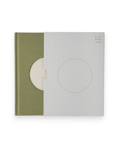 A Wilde House Paper hardcover journal, moss-coloured with a large circular cutout in the center is semi exposed from its grey coloured case.