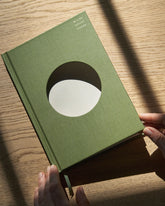 A Wilde House Paper hardcover journal, moss-coloured with a large circular cutout in the center is held in two hands with a light wood surface as the full background. 
