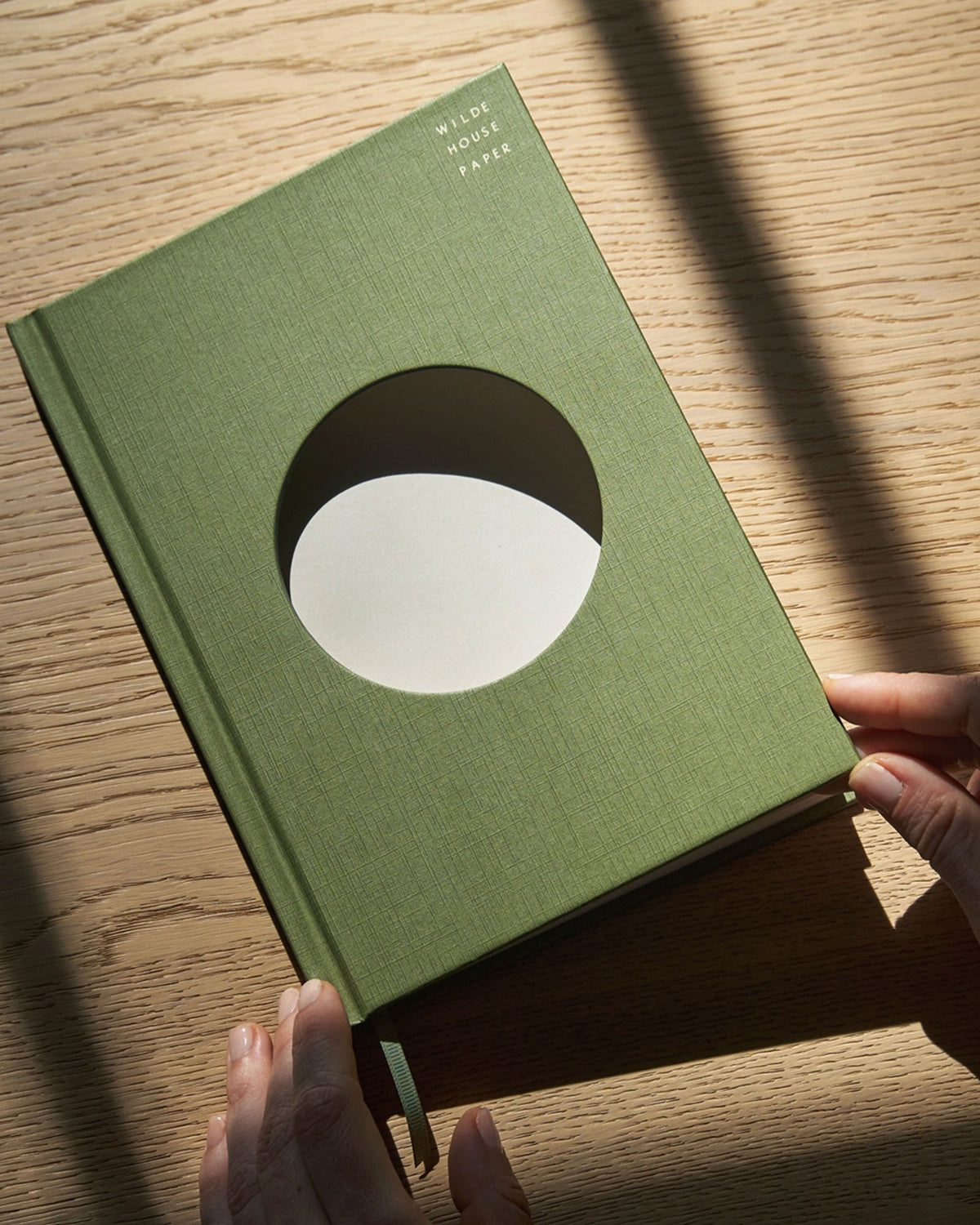 A Wilde House Paper hardcover journal, moss-coloured with a large circular cutout in the center is held in two hands with a light wood surface as the full background. 