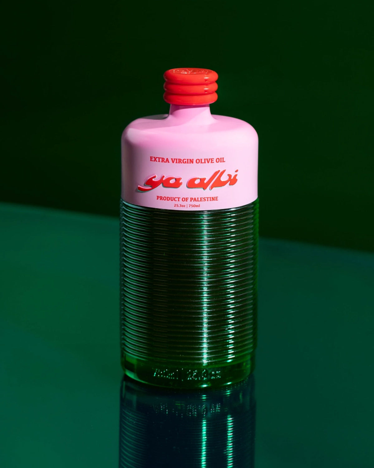 A bottle of Ya Albi extra virgin olive oil, featuring a pink upper label and cap with a ribbed green glass body, set against a dark green background with a reflection below.