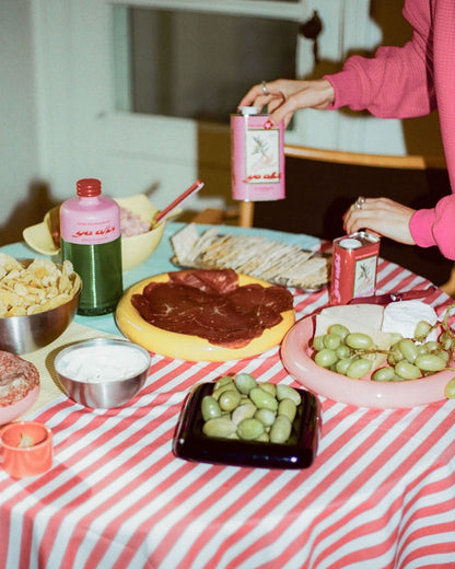 A styled food table scene with a red-and-white striped tablecloth, platters of sliced meat, green olives, grapes, chips, and Ya Albi&