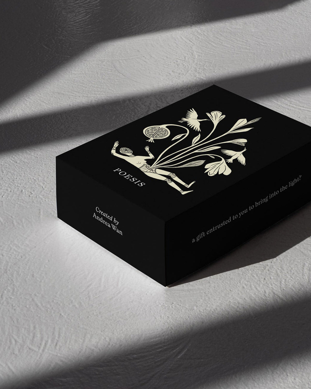 Black rectangular box with an off-white, hand drawn illustration of a body laying horizontally, the head is drawn as a cut piece of fruit and tall stemmed flowers are blooming from the torso. An open-winged bird is on each side of the body bouquet. The word POESIS is written under the illustration. Box is on a textured white table surface with dramatic shadows. 