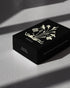 Black rectangular box with an off-white, hand drawn illustration of a body laying horizontally, the head is drawn as a cut piece of fruit and tall stemmed flowers are blooming from the torso. An open-winged bird is on each side of the body bouquet. The word POESIS is written under the illustration. Box is on a textured white table surface with dramatic shadows. 